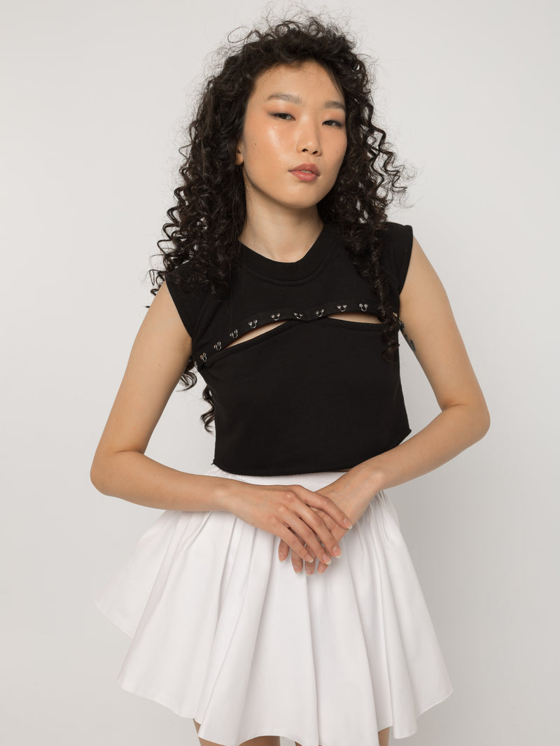 Model wearing Girl Code black pierced cutout crop top with a white pleated skirt